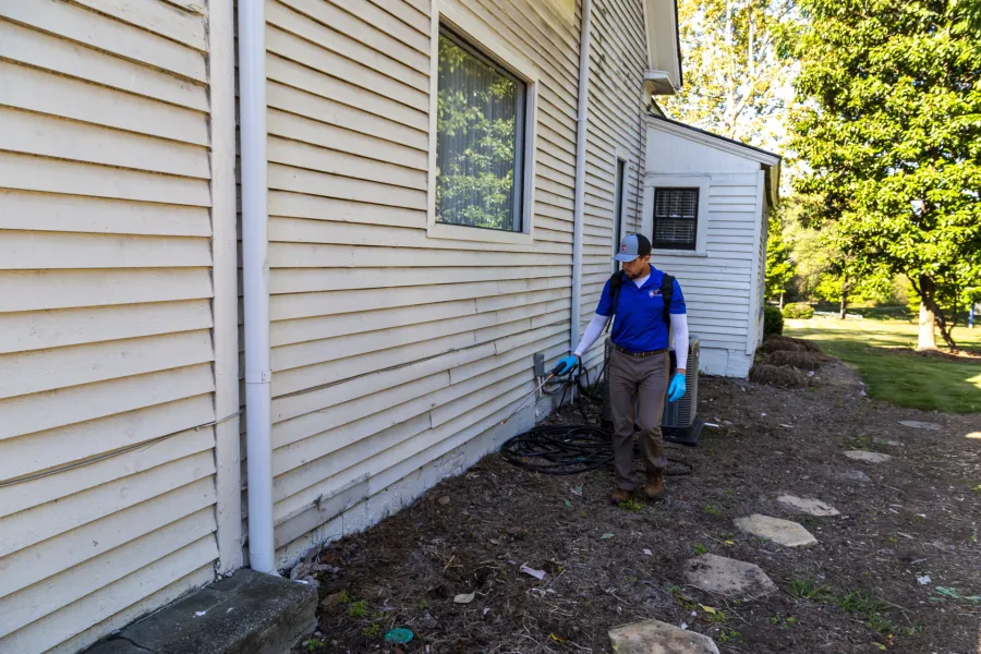 Pest control technician sprays exterior of beige house siding using spray equipment on sunny day