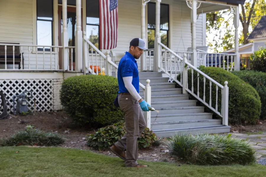 Man in blue shirt and cap inspecting lawn with handheld device in front of a white house with American flag.