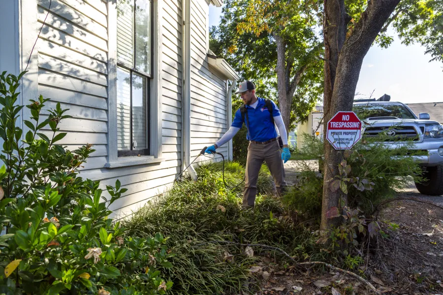 Technician spraying pesticide on exterior house wall near garden plants with 'No Trespassing' sign on tree.