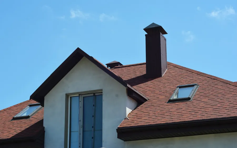 Modern house roof with red shingles, skylights, and a prominent brown chimney under clear blue sky.