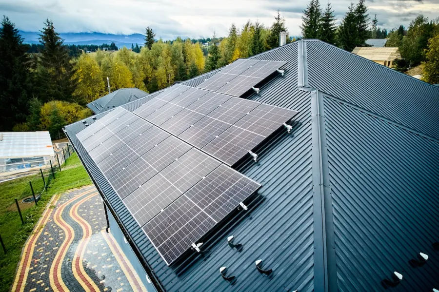 Solar panels installed on a dark metal roof with forest and mountains in the background under a cloudy sky
