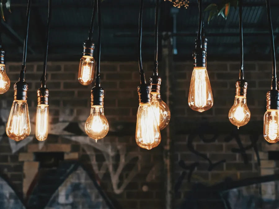 Multiple vintage Edison light bulbs hanging from cords against a dark brick wall with graffiti.