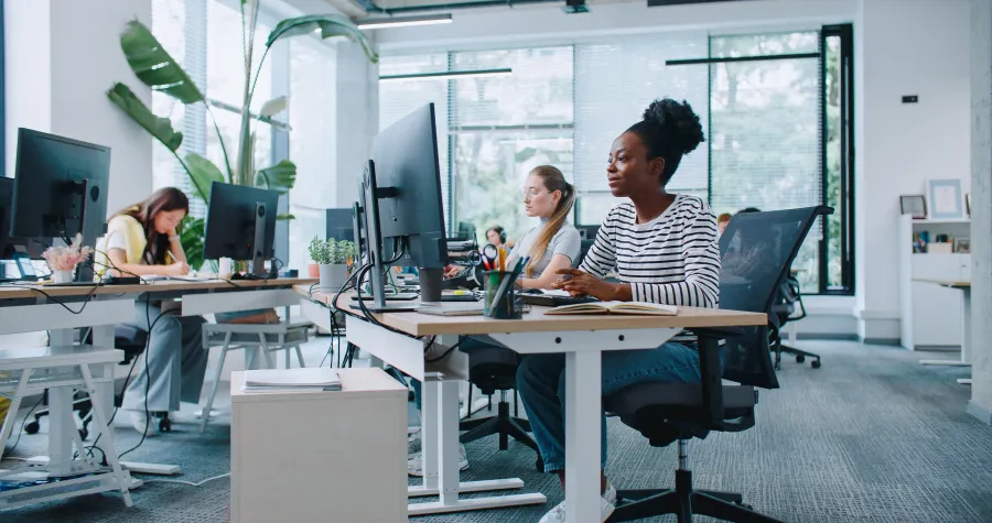 Two women working at desks in a modern office with plants and bright light streaming through windows.