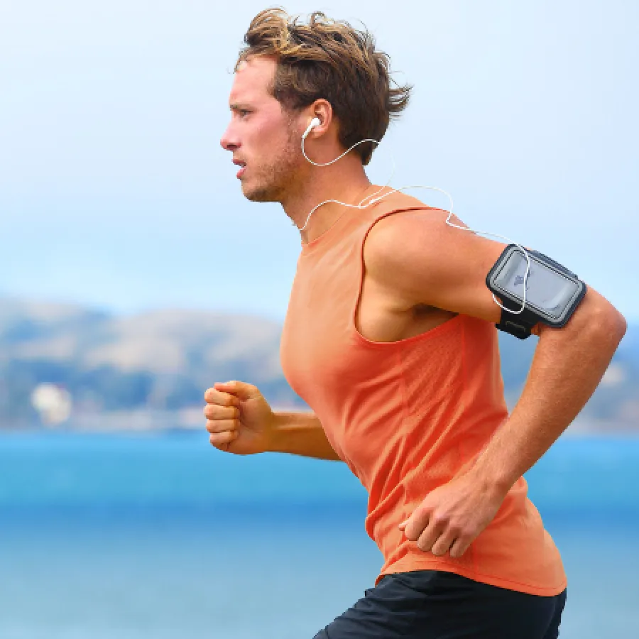 Runner in an orange sleeveless shirt and black shorts jogging by the water with earphones and armband phone holder.