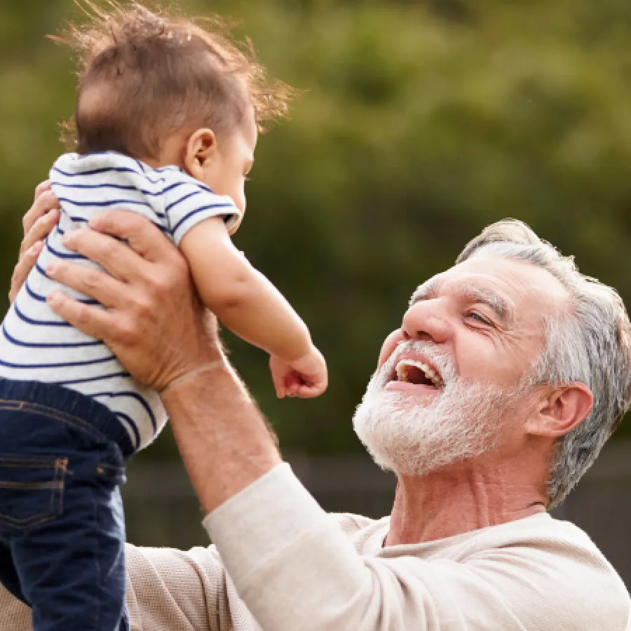 Happy grandfather lifting and playing with baby boy outside in a green garden on a sunny day