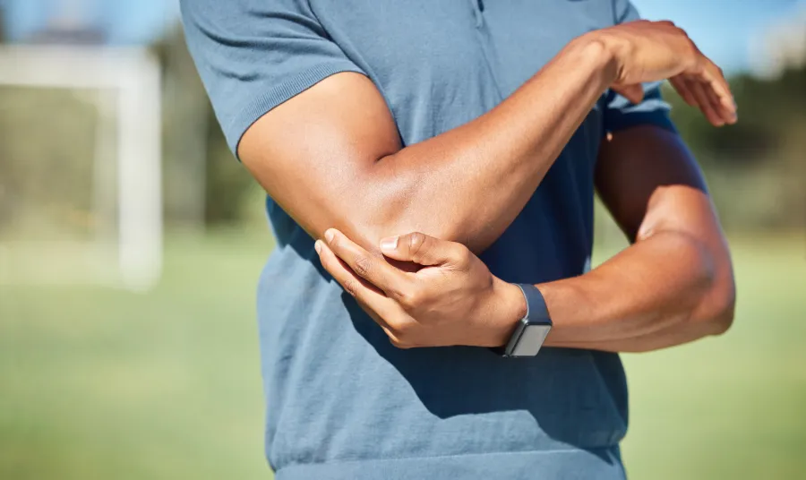 Man in blue shirt holding his elbow outdoors indicating elbow pain or injury during sports activity
