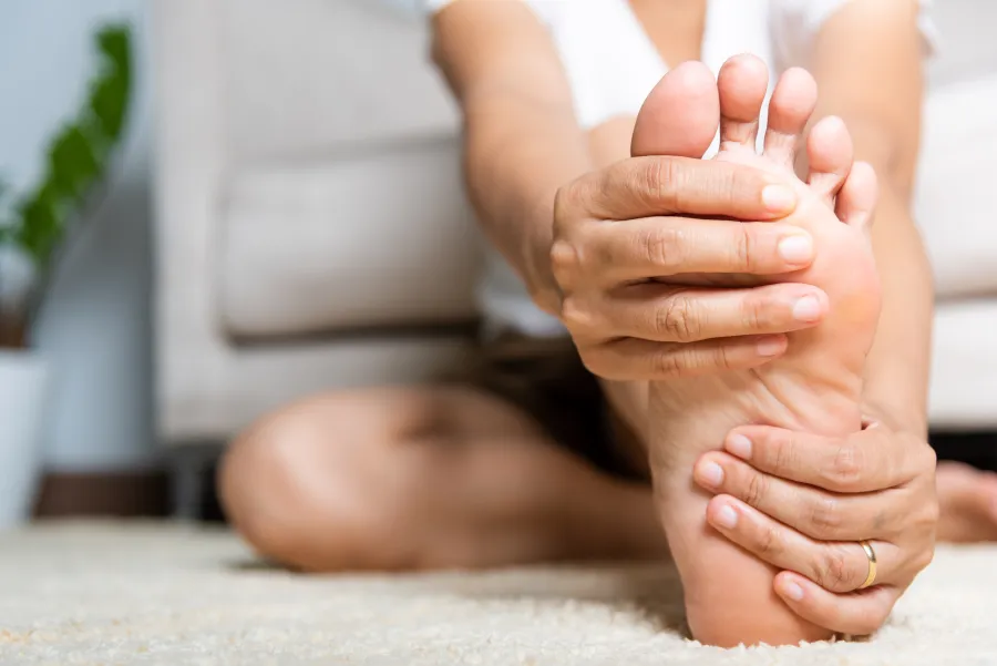 Person sitting on floor holding and stretching their bare foot with both hands indoors on carpet.