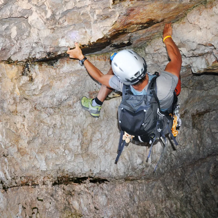 Male rock climber wearing helmet and backpack ascends challenging overhang in rocky cave environment.