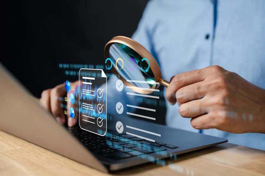 Person using magnifying glass to review digital document checklist on a laptop keyboard with holographic data interface.