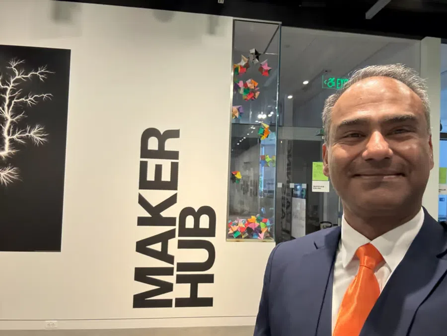 Man in a suit with an orange tie smiling inside a Maker Hub with colorful origami and creative artwork.