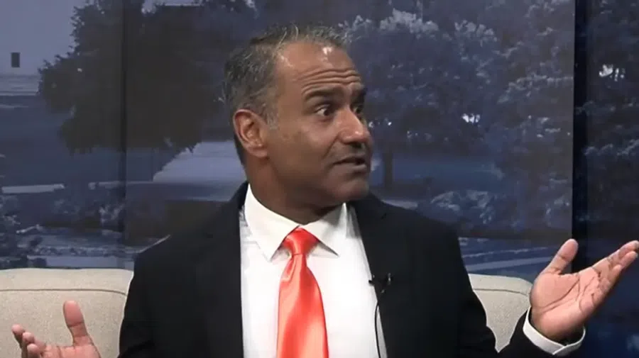Man in black suit and bright orange tie gestures with hands while speaking on a talk show set with dark backdrop.