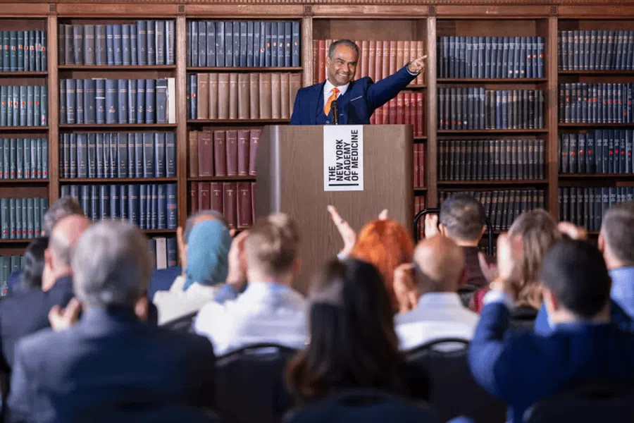 Speaker in a suit addressing an applauding audience at a medical academy event with bookshelves in the background