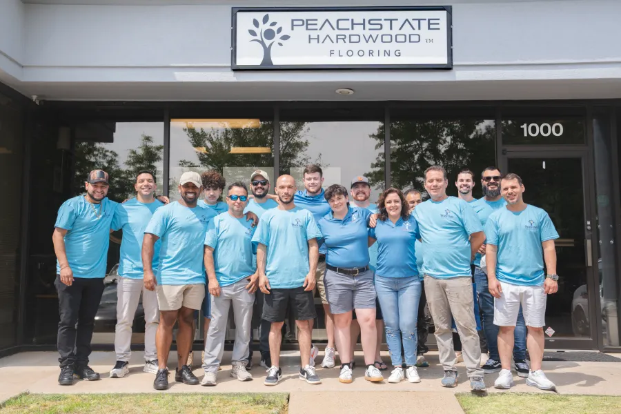 Team of employees in matching blue shirts standing outside Peachstate Hardwood Flooring storefront under the company sign.
