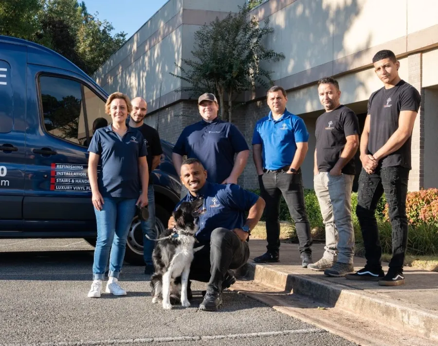 Group of seven workers standing outside a building near a company van, one kneeling with a black and white dog.
