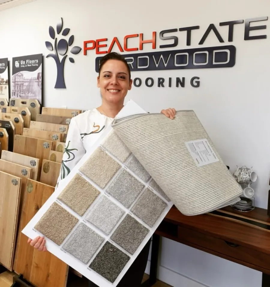 Smiling woman holding carpet and hardwood flooring samples inside a flooring showroom with Peach State Hardwood logo.
