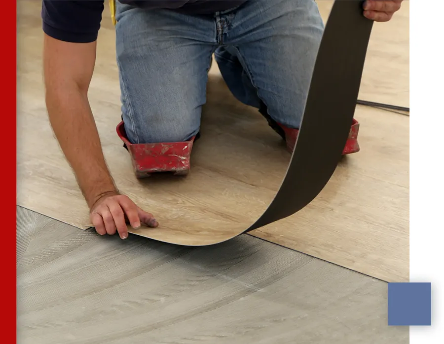 Person installing wood-look vinyl flooring on a gray subfloor, wearing knee pads and jeans, hands positioning the flooring.