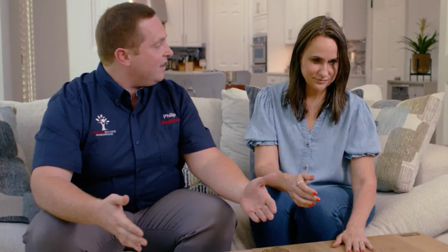 Man and woman sitting on a couch discussing while gesturing over a wooden table in a living room.