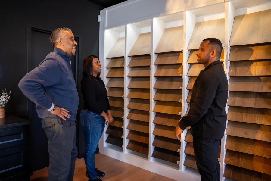 Three people discussing wood flooring samples displayed on a wall in a showroom setting.