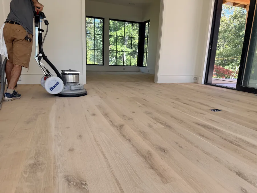 Person using a Bona floor cleaning machine on light wooden flooring in a bright room with large windows.