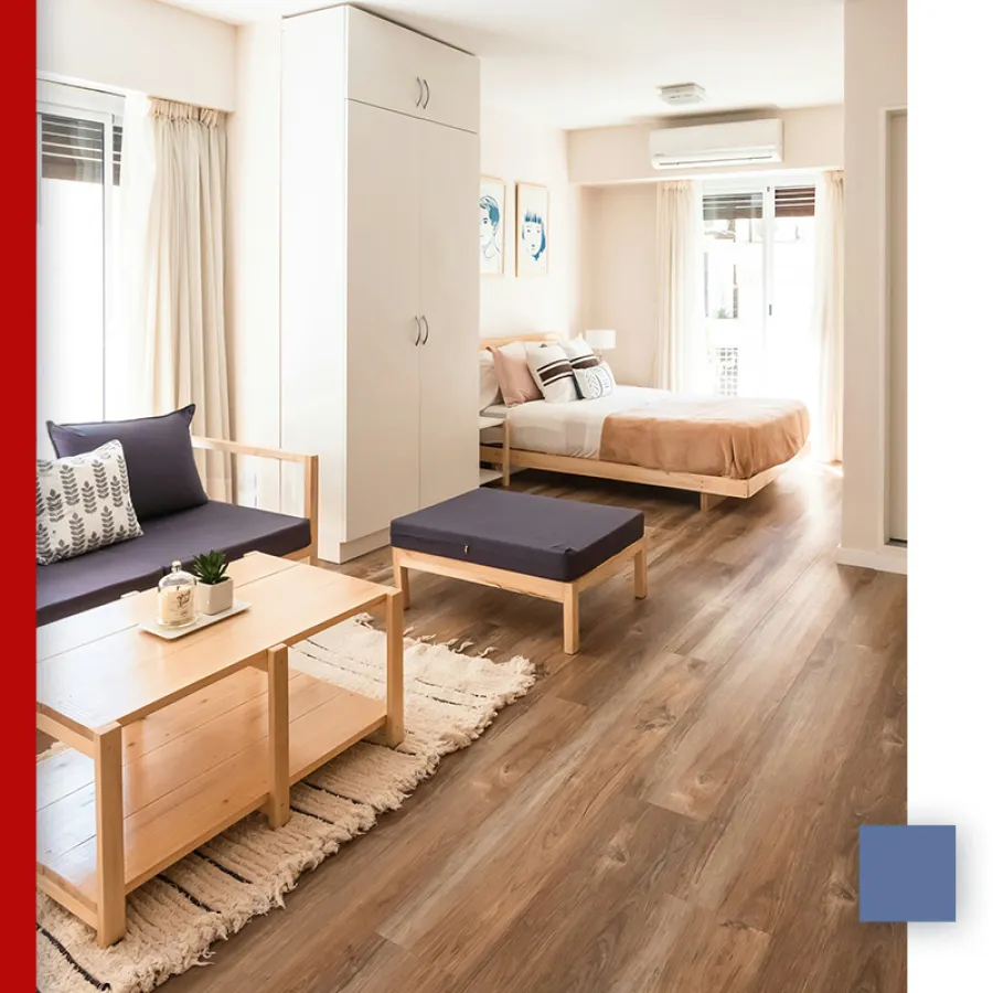 Bright modern studio apartment with wooden floors, beige bed, navy cushions, and light wooden furniture.