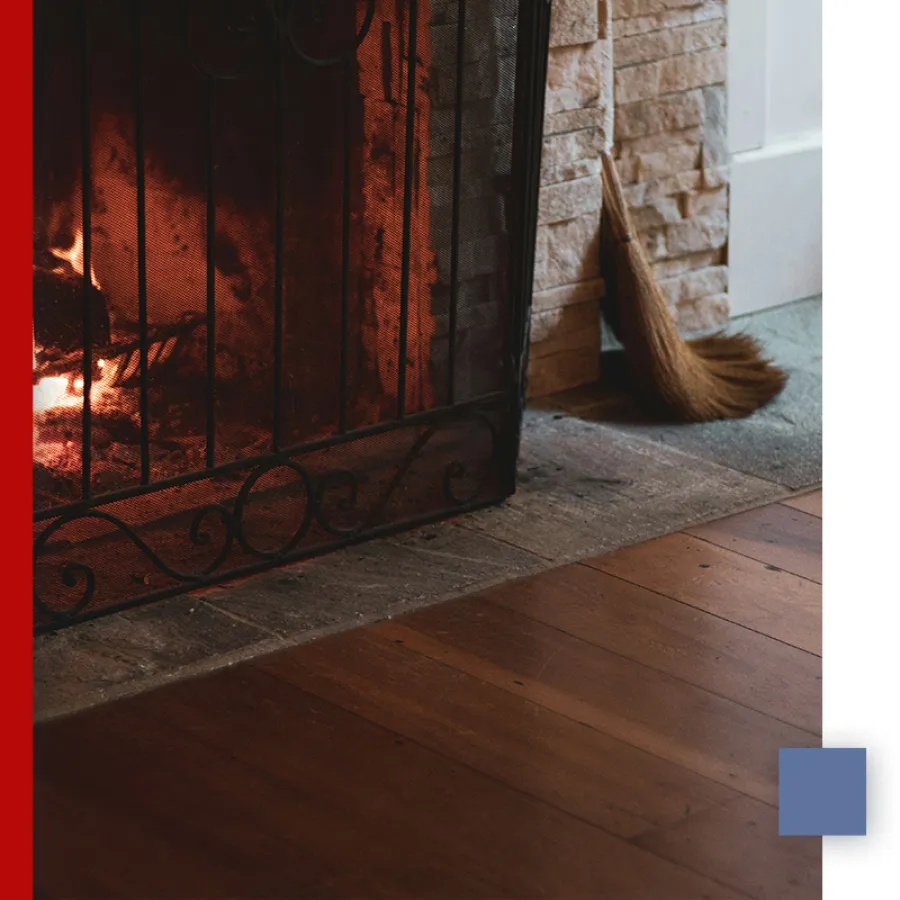 Cozy fireplace with metal screen, lit fire, stone wall, wooden floor, and straw broom resting nearby.