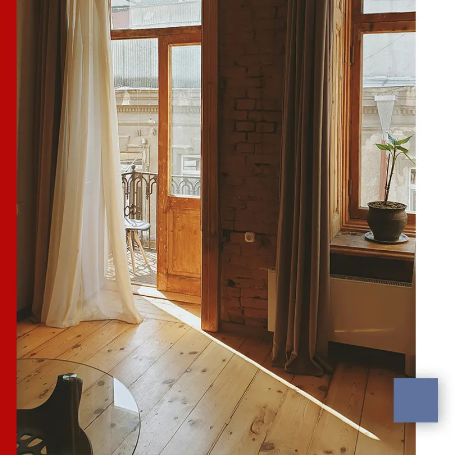 Sunlit cozy room with wooden floor, open door to balcony, sheer and dark curtains, and potted plant on window sill