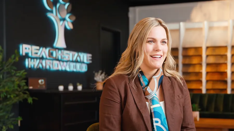 Smiling woman in business attire sitting in a hardwood showroom with sample boards and PEACH STATE HARDWOOD logo.