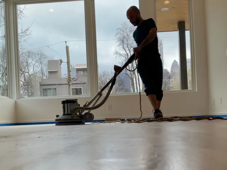 Man using floor polishing machine on wooden floor inside a bright room with large windows.