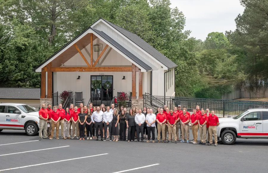 Large team of PestNation employees in red and black uniforms standing in front of white office building with company trucks.