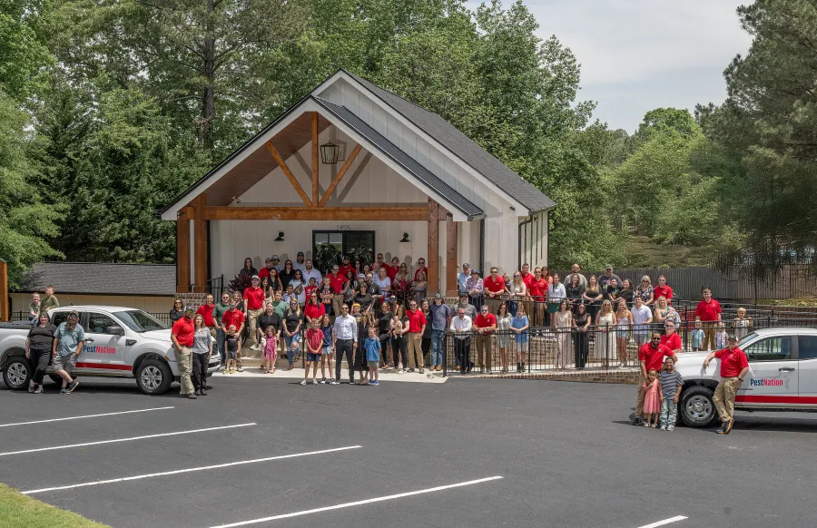 Large group of people posing outside a modern building with two Pest Nation trucks in a parking lot surrounded by trees