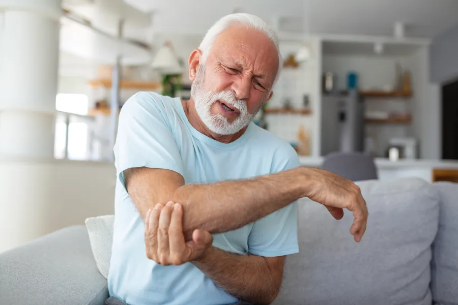 Elderly man in light blue shirt sitting on couch, holding elbow with a pained expression in a living room.