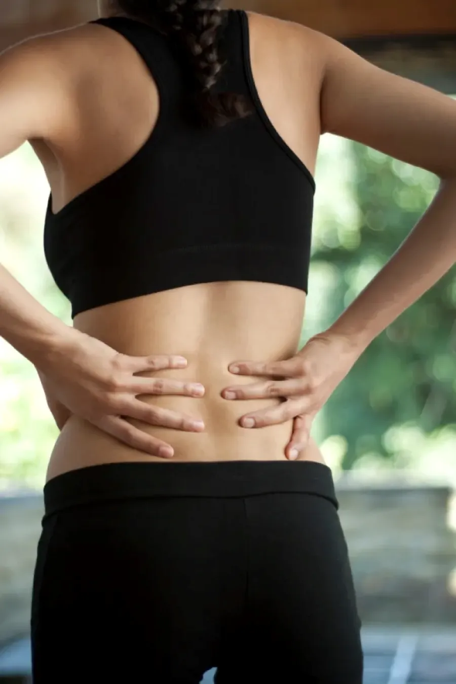Woman in black sportswear holding lower back in pain, indicating backache or muscle strain indoors.