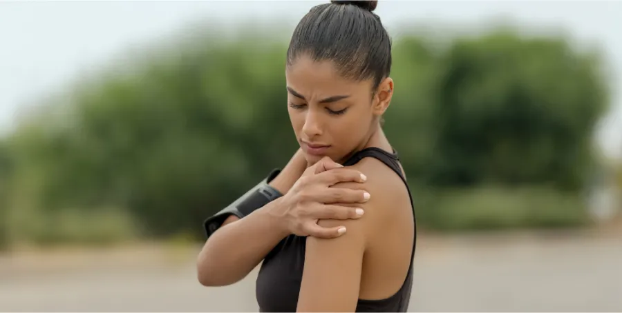 Woman in athletic wear holding her shoulder in pain outdoors, illustrating preparing for shoulder arthroscopy surgery.