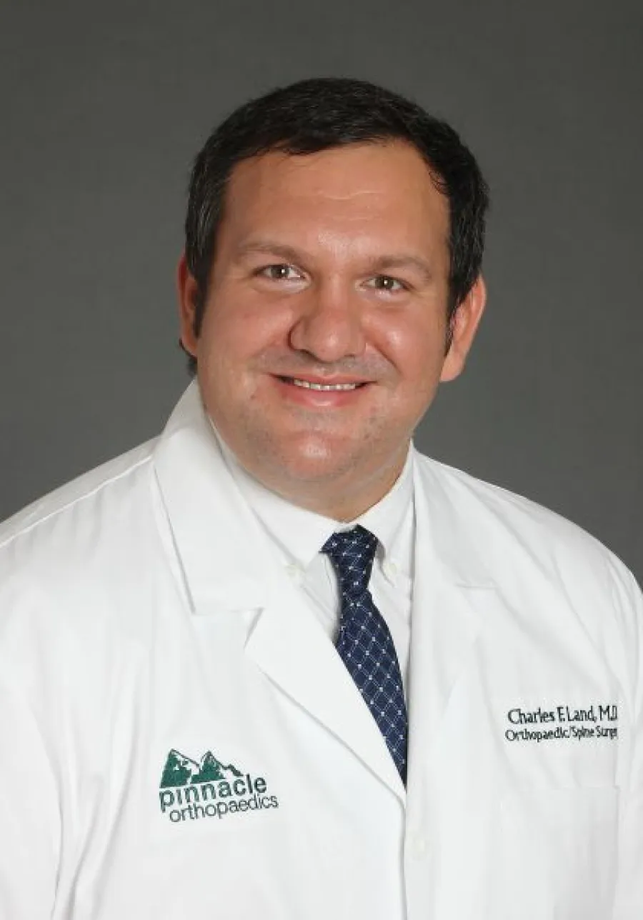 Smiling male doctor in white coat with Pinnacle Orthopaedics logo and blue tie on gray background.
