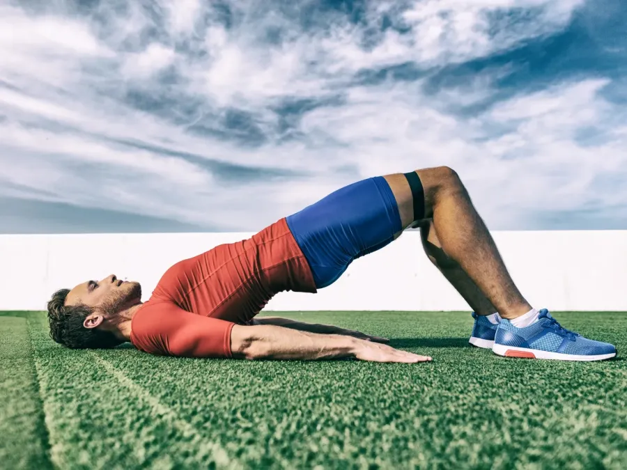 Man in red shirt and blue shorts doing a bridge exercise on green grass under blue cloudy sky