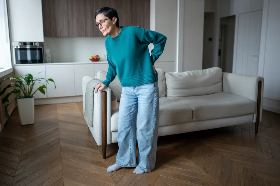 Woman in teal sweater and jeans holding her lower back, standing in a modern living room with a beige sofa.