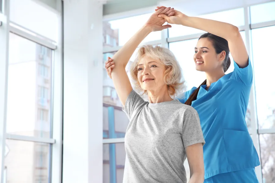 Senior woman stretching arms with physical therapist assistance in bright modern rehab center.