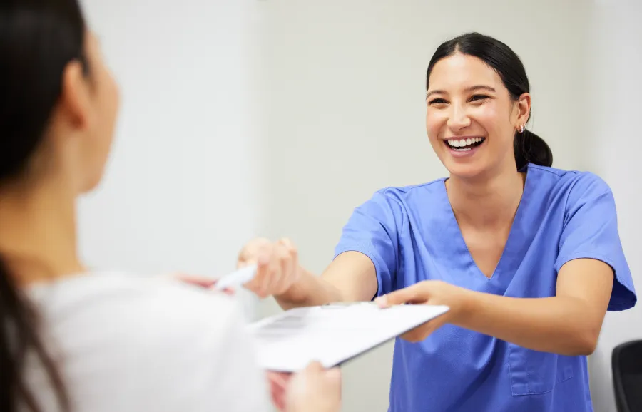 Smiling healthcare worker in blue scrubs handing clipboard to patient in bright medical office.