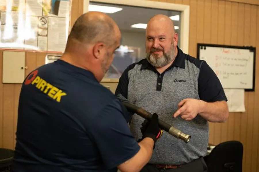 Two men in work uniforms inspecting a hose assembly in an industrial setting with wooden walls.