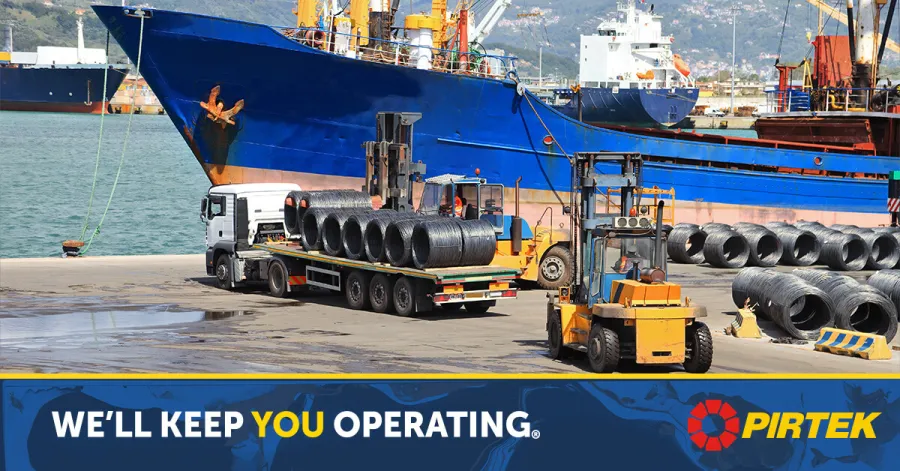 Forklift loading steel coils onto a truck at a busy port with docked cargo ships in the background