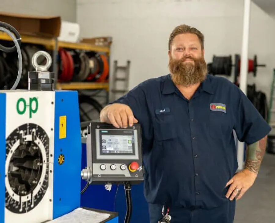 Bearded technician in navy uniform standing next to industrial machine control panel in workshop.