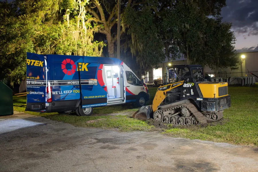 Mobile service van and compact tracked excavator parked on grassy area at dusk with trees and building in background