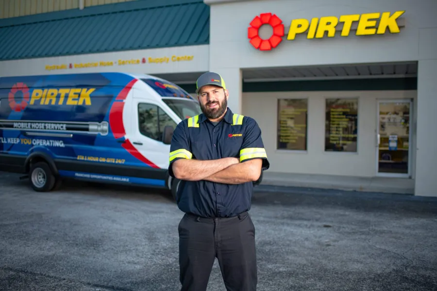 Pirtek technician stands confidently outside service center with branded mobile hose service van in background.