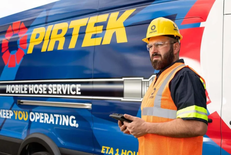 PIRTEK worker in safety gear stands by branded van offering mobile hose service with tablet in hand.
