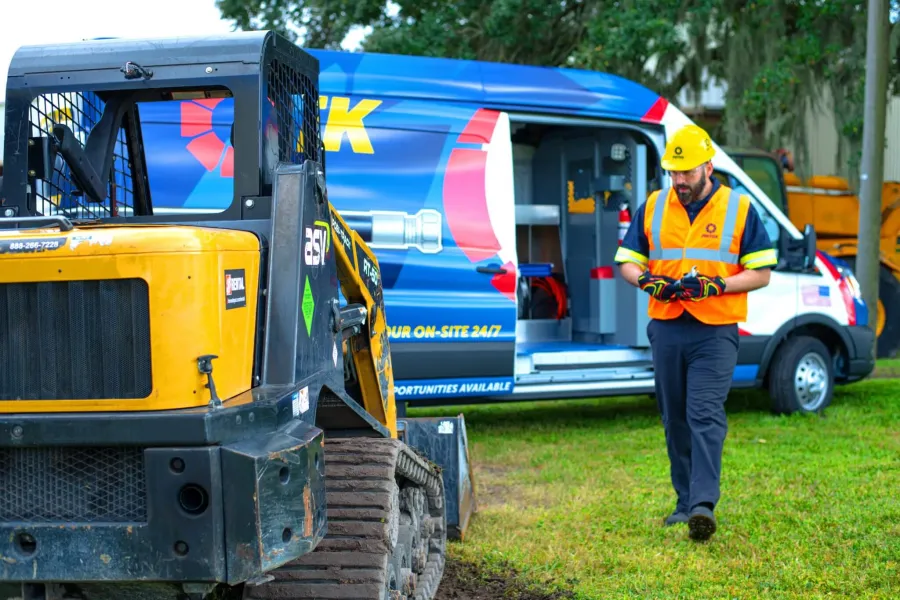 Construction worker in safety gear using device near heavy machinery and service van on grassy site