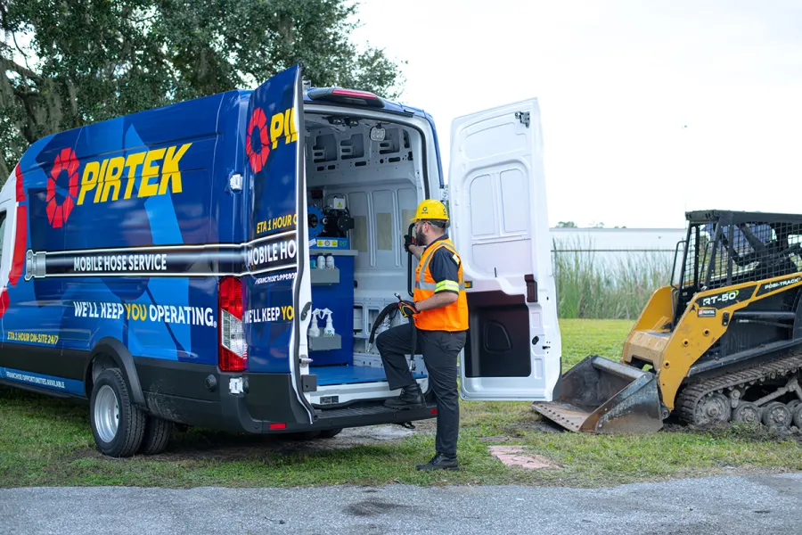 Pirtek mobile hose service van with technician in safety gear working beside a construction site with skid steer loader.