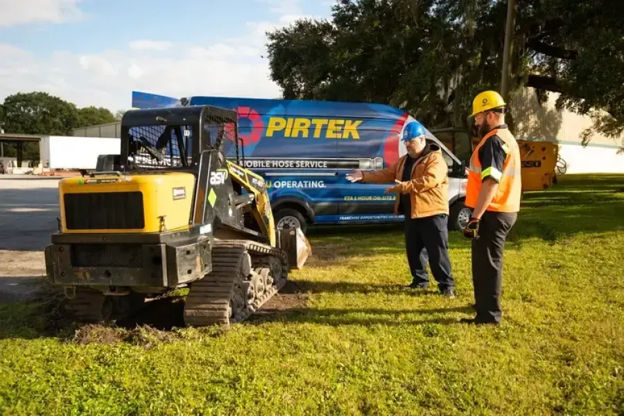 Two construction workers inspecting a compact tracked loader near a PIRTEK service van on grassy ground.