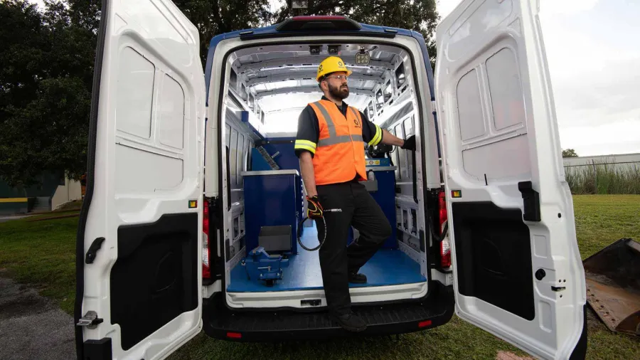 Construction worker in safety gear standing inside an open white service van outdoors on a grassy area.