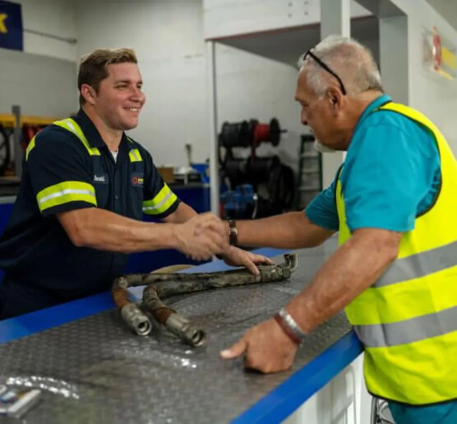 Two men shaking hands over a workbench in an industrial workshop setting with tools and pipes