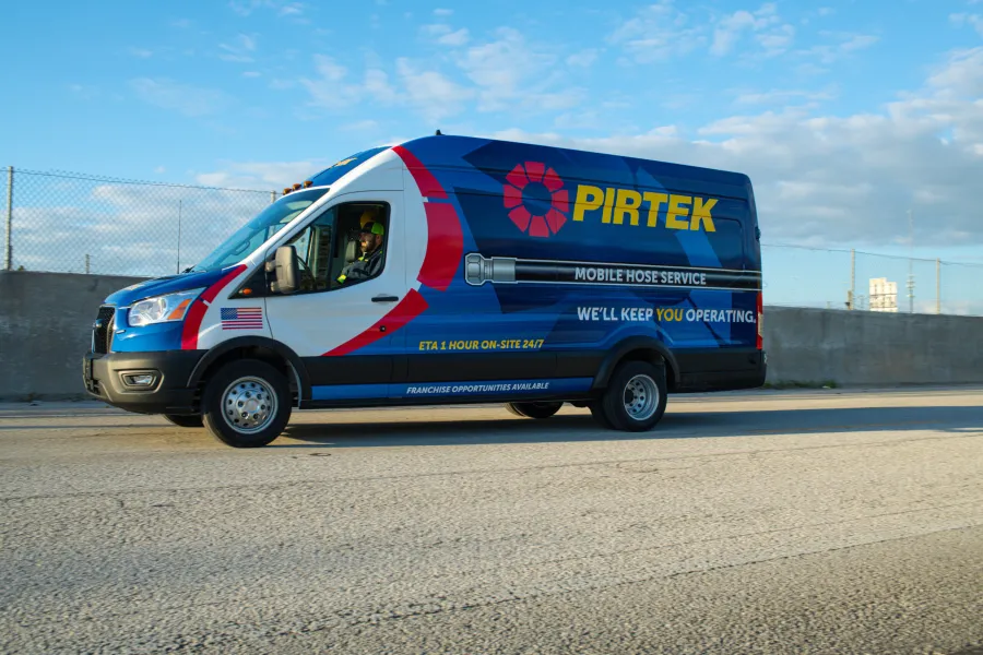 Blue and white Pirtek mobile hose service van driving on a road with clear sky background.